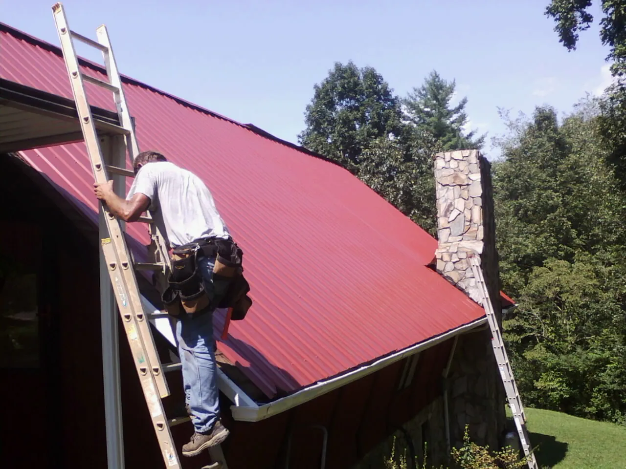 Roofer on a ladder beside a red metal roof and stone chimney — Soffit & Fascia Repair by Roofing Brothers in Great Valley